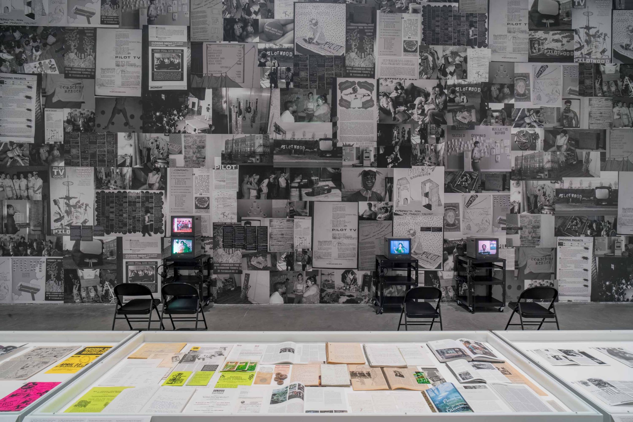 City in a Garden: Queer Art and Activism exhibit in Chicago that features a collage of Black and white images on a wall with a table of previous flyers advertising queer events of Chicago's past.